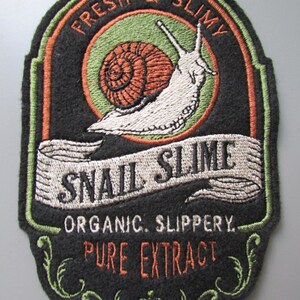 Könnte beinhalten: Ein bestickter Aufnäher mit schwarzem Hintergrund, der eine weiße Schnecke mit orangefarbenem Gehäuse zeigt. Der Text lautet FRESH & SLIMY, SNAIL SLIME, ORGANIC. SLIPPERY. und PURE EXTRACT. Das Design enthält grüne kreisförmige Akzente und Ornamente.