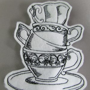 May include: Black and white embroidered design of a teacup and saucer with a teapot lid on top. The design is on a white felt background.