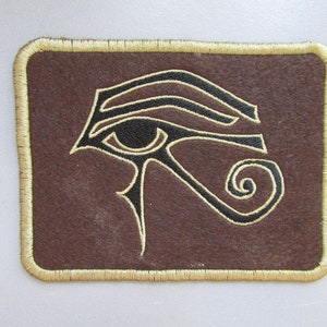 May include: A brown embroidered patch featuring a black eye of Horus design. The patch has a gold stitched border.
