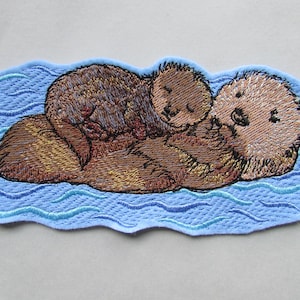 May include: Embroidered patch depicting two brown sea otters cuddling in blue water. The larger otter is on its back with its paws wrapped around the smaller otter. The smaller otter is curled up on the larger otter's chest.