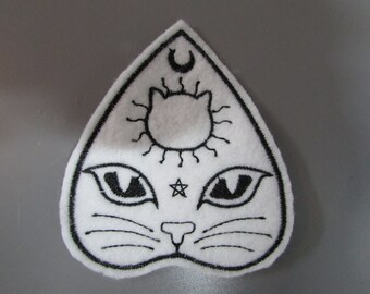 Puede incluir: Parche de fieltro blanco en forma de familiar de bruja con detalles bordados en negro. El parche presenta la cara de un gato con una luna creciente, el sol y una estrella de cinco puntas.