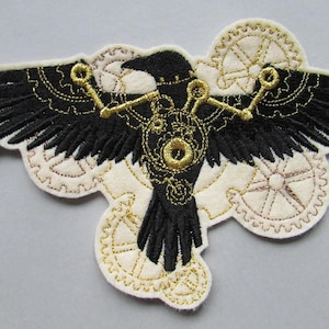 Könnte beinhalten: Eine schwarze gestickte Eisenkrähe mit goldenen Zahnrädern und Zahnrädern auf weißem Hintergrund.