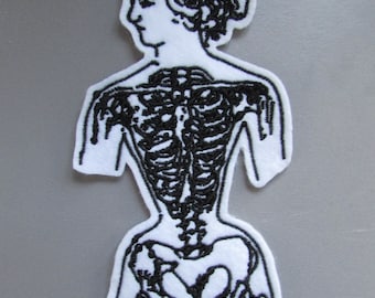 May include: White embroidered patch of a skeleton with a human-like face and hair. The skeleton is wearing a corset and has a detailed rib cage and pelvic bone.