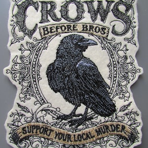 May include: An off-white embroidered patch featuring a black crow centered within an ornate oval frame. The text reads "CROWS BEFORE BROS" at the top and "SUPPORT YOUR LOCAL MURDER" on a banner at the bottom, all rendered in black and gold thread.