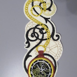 May include: An embroidered patch featuring a pocket watch design with a gold chain and black filigree on an off-white background. The watch has a black face with gold detailing and a red top.
