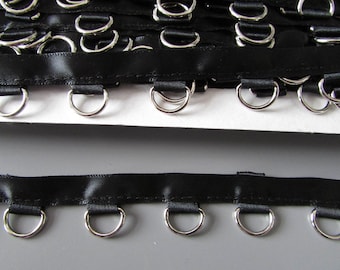 May include: Close-up of black satin ribbon with evenly spaced silver metal D-rings. The ribbon is sewn to the rings, creating a decorative or functional trim. The background is a neutral grey and white.