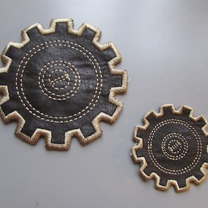May include: Two brown fabric gear-shaped coasters with gold stitching. The larger coaster is in the background and the smaller coaster is in the foreground.