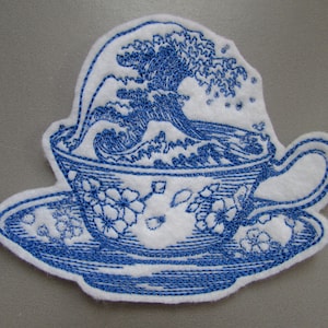 May include: Blue embroidered patch depicting a teacup with a wave crashing over the rim. The teacup and saucer are decorated with a floral pattern.