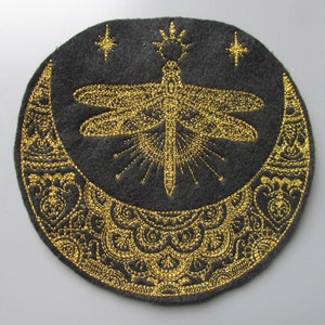 以下が含まれることがあります： 金色の刺繍が施された黒いフェルトのパッチ。トンボ、三日月、星がデザインされています。トンボは中央にあり、その背後には太陽の光が放射状に広がっています。三日月は装飾的なパターンで縁取られています。