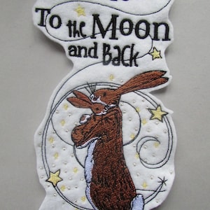 Könnte beinhalten: Gestickter Aufnäher mit einem braunen Kaninchen, das ein kleineres Kaninchen hält, umgeben von einem silbernen Wirbel mit gelben Sternen. Der Text "I Love You To the Moon and Back" ist in Schwarz gestickt.
