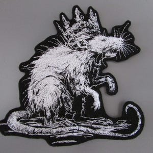 May include: Embroidered patch in black and white, showing a rat wearing a crown. The design uses white thread to create a detailed image of the rat on a black backing. The patch has an unusual, non-uniform shape.