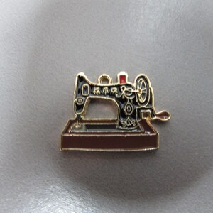 Pode incluir: Encanto de metal dourado em forma de máquina de costura preta com um detalhe vermelho. A máquina tem texto japonês no lado.