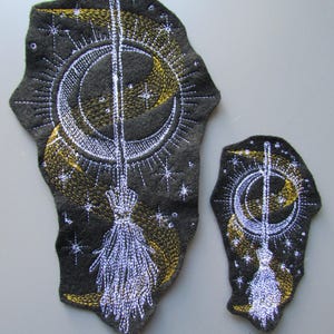 Könnte beinhalten: Zwei schwarze Filzaufnäher mit gestickten Motiven eines Besenstiels, der vertikal über einem Halbmond positioniert ist. Die Stickerei verwendet metallisch goldenen Faden für den Mond und silbernen Faden für den Besen und die Sternendetails.