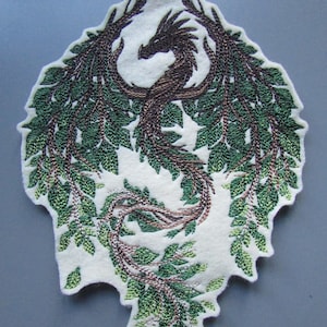 Könnte beinhalten: Gestickter Aufnäher mit einem grünen und braunen Drachen, dessen Flügel aus Blättern bestehen. Der Drache ist zu einem Kreis zusammengerollt, mit dem Kopf zum Betrachter gewandt. Der Aufnäher ist aus Filz und hat einen weißen Hintergrund.