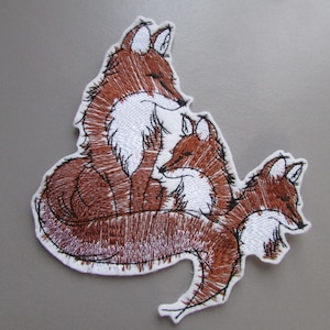 May include: Embroidered patch featuring three brown foxes curled up together. The foxes are outlined in black thread and have white accents on their fur.