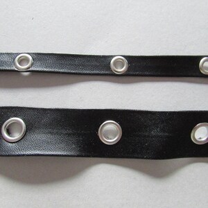 May include: Two black leather strips, each featuring a series of silver eyelets. The eyelets are circular and evenly spaced along the length of the strips. The leather has a smooth texture, and the eyelets are securely fixed.