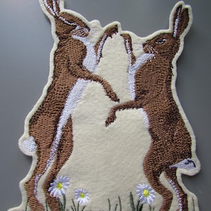 May include: An embroidered fabric patch featuring two brown hares standing on their hind legs facing each other. The hares have white underbellies and tails. Below them are three small white daisies with yellow centers and green stems on a patch of grass.