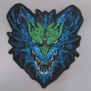 May include: Embroidered patch featuring a stylized dragon's head. The design showcases a black background with a detailed dragon face in shades of blue and green, with white fangs and eyes. The patch has a textured appearance.