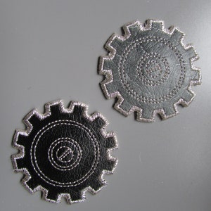 May include: Two black leather gear-shaped coasters with silver stitching. The coasters are on a gray background.