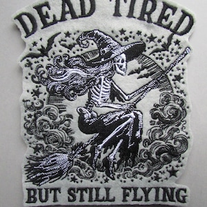 May include: Black and white embroidered patch featuring a skeleton witch flying on a broomstick. The text reads "Dead Tired But Still Flying".