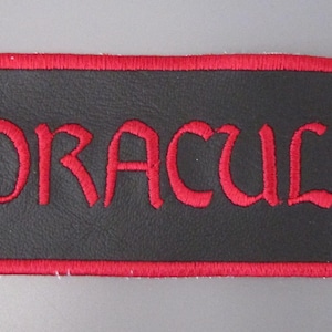 May include: A rectangular patch with a black background and a red border. The word "DRACULA" is embroidered in red, along with a red bat-like shape on the left. The text "BRAM STOKER" is embroidered in red on the right.