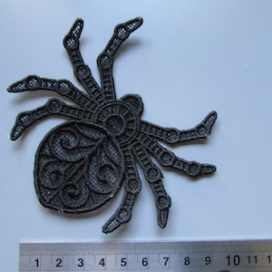 May include: Black lace spider applique with detailed design. The spider features eight legs and a rounded body. The applique is approximately 10 cm wide, ideal for Halloween or craft projects.