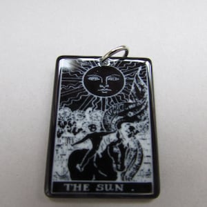 May include: Black and white tarot card pendant with a silver bail. The card depicts a sun with a face, a figure riding a horse, and the text "THE SUN."