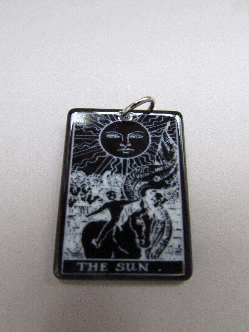 May include: Black and white tarot card pendant with a silver bail. The card depicts a sun with a face, a figure riding a horse, and the text "THE SUN."
