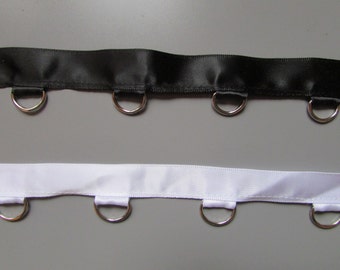 May include: Two strips of satin ribbon, one black and one white, each with silver metal D-rings attached. The ribbons are displayed against a grey background, showcasing the contrasting colours and textures of the materials.