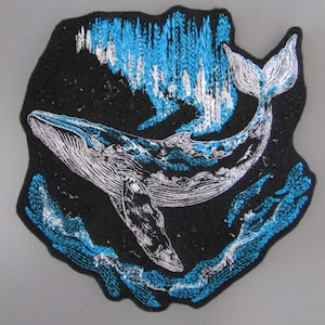 May include: Embroidered patch depicting a whale in the sea. The whale is white and blue, set against a black backdrop with blue and silver details. The patch has an unusual, irregular shape.