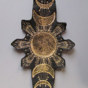 May include: A black fabric patch with gold embroidery depicting a sun with rays and three crescent moons. The design is reminiscent of celestial imagery.