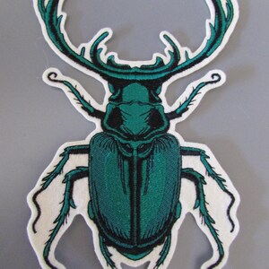 May include: Embroidered patch featuring a detailed teal beetle design with prominent antennae and legs. The insect is set against a white background with a stitched border, creating a striking contrast. The patch is likely for clothing or accessories.