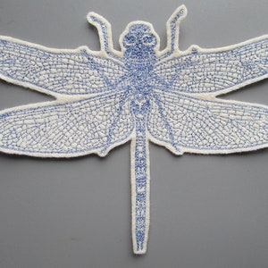 May include: A blue embroidered dragonfly applique with a white background. The dragonfly has detailed stitching on its wings and body.