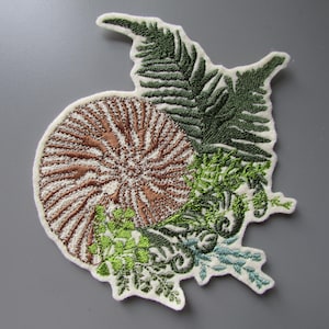 May include: An embroidered patch featuring a brown nautilus shell design layered with dark green fern fronds and light green foliage. The entire piece is outlined in white thread, creating a distinct shape against the background.