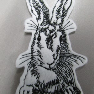 May include: Black and white embroidered patch of a rabbit with long ears. The rabbit is sitting with its front paws in front of it.