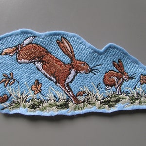 May include: Embroidered patch depicting two brown rabbits hopping through a field of green grass and blue sky. The larger rabbit is leaping in the air, while the smaller rabbit is hopping on the ground.