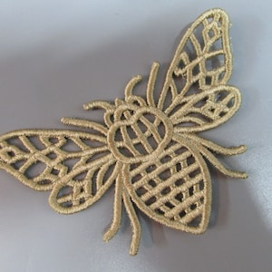 May include: A gold-coloured embroidered bee applique with a detailed, intricate design. The bee has a large, round body and delicate wings.