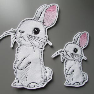 May include: Two white rabbit patches with black embroidered outlines and details. Each rabbit is standing on its hind legs with large dark eyes, long whiskers, and pink-tinted inner ears. The patches are shown in two different sizes.