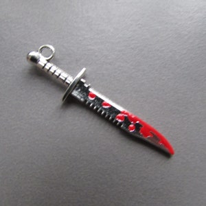 May include: A silver-coloured knife charm with a red blood splatter design. The knife has a serrated blade and a loop at the top for hanging.