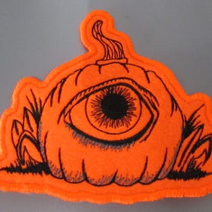 May include: Orange felt patch depicting a pumpkin with a large eye and black outline. The eye has a detailed iris and pupil. The patch has a black outline and is surrounded by black grass.