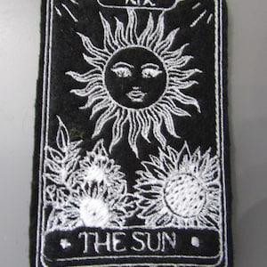 May include: Black felt patch with white embroidery of the tarot card The Sun. The card number XIX is in the top left corner. The sun is depicted as a stylized face with rays emanating from it. Two sunflowers are below the sun.