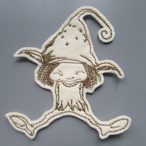 May include: A white felt appliqué of a smiling gnome wearing a pointed hat with a curled brim. The gnome has brown hair and is wearing a white shirt with a brown embroidered design. The gnome is running with its arms outstretched.