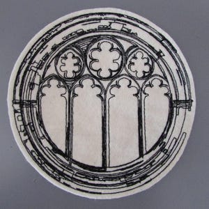 Puede incluir: Un posavasos redondo, blanquecino, con una ilustración negra detallada de una ventana de catedral. El diseño presenta ventanas arqueadas y patrones circulares, creando una estética gótica. El posavasos es probablemente de fieltro.