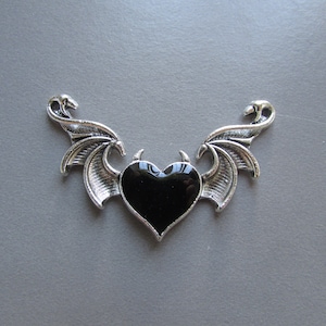 Peut inclure: Pendentif en métal argenté avec un cœur noir, de petites cornes et des ailes de chauve-souris stylisées. Le cœur est brillant et les ailes ont des détails de plumes. Pour bijoux ou travaux manuels.