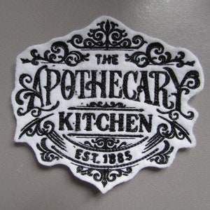 Könnte beinhalten: Weißer Filz-Patch mit schwarzem gesticktem Text, der "The Apothecary Kitchen Est. 1885" lautet. Der Text ist von dekorativen Schnörkeln umgeben.
