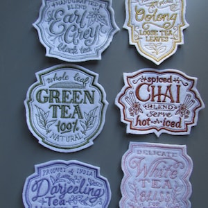 Peut inclure: Six étiquettes brodées pour sachets de thé avec différentes variétés de thé. Les étiquettes sont blanches avec des broderies et du texte colorés. Les étiquettes comprennent Earl Grey, Thé vert, Thé de Darjeeling, Thé blanc Bliss, Chai et Oolong.