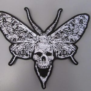 May include: A black and white embroidered patch featuring a moth with a skull design. The moth has detailed white wings and a skull head. The patch has a black border and is suitable for jackets, bags, or other fabric items.