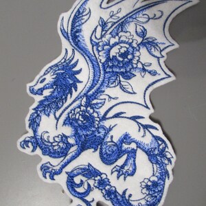 May include: Embroidered patch depicting a blue dragon with floral details on a white backing. The dragon design includes wings and is surrounded by flowers and foliage. Suitable for application on clothing or accessories.
