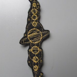 Op de afbeelding: Een zwarte viltpatch met gouden borduurwerk dat een gestileerd zonnestelsel met planeten en ringen voorstelt.
