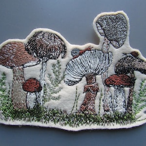 May include: Embroidered patch featuring a group of mushrooms in various colours and shapes, including brown, white, and red, with green foliage surrounding them.